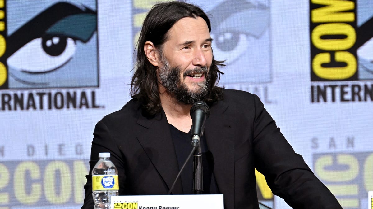 Keanu Reeves in a black suit, smiling as he speaks at a panel with Comic-Con International backsplash behind him.