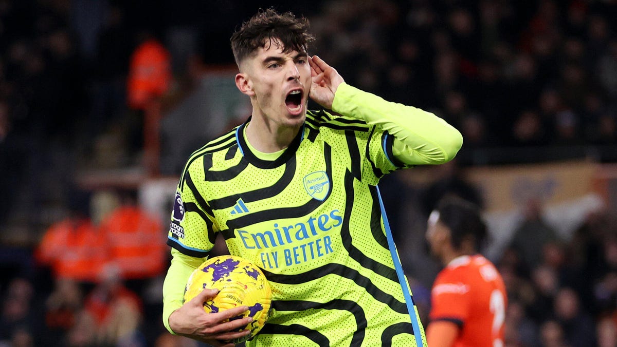 Arsenal forward Kai Havertz celebrating, holding a ball under his right arm while cupping his right ear with his right hand.