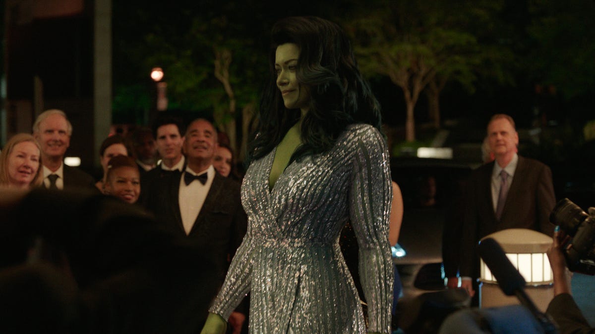 She-Hulk, a woman with green skin and superpowers, struts into a black tie event wearing a sparkling silver dress.