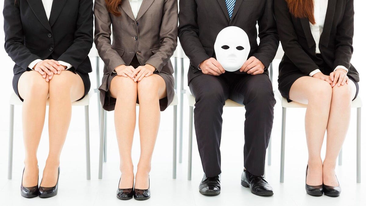 Four people in business attire sit in a row, framed from the shoulders down, with one holding a suspicious white mask.