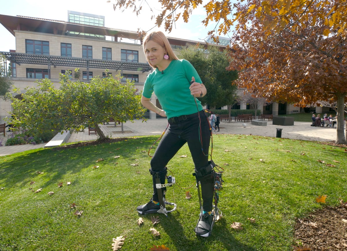 Uma mulher fica do lado de fora na grama usando duas botas de exoesqueleto robótico