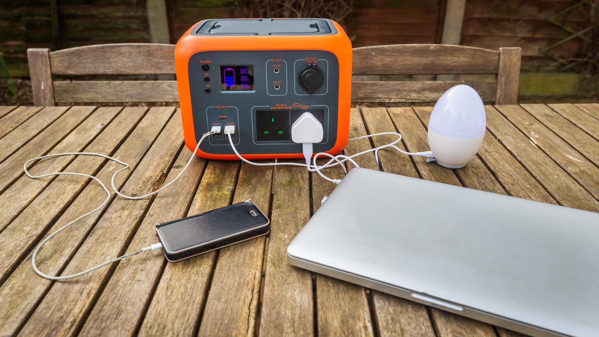 Portable power station solar electricity generator outdoors on wooden table with laptop, mobile phone and light electronic devices charging. Wireless charging lithium battery backup for use off grid.