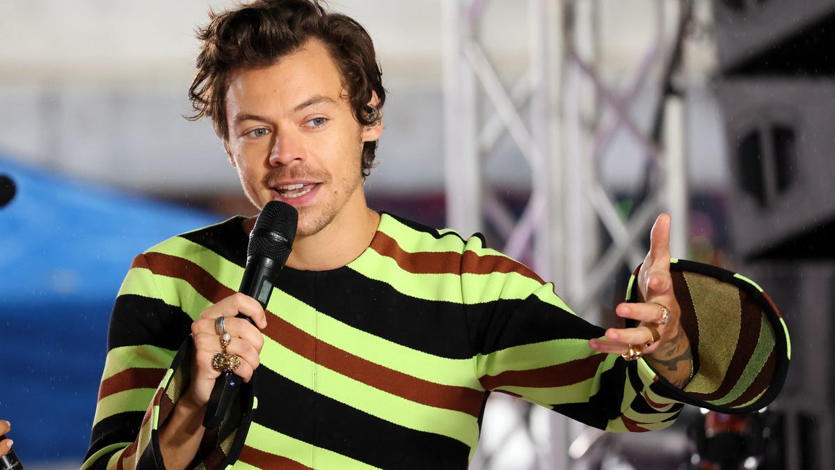 Harry Styles wears a striped shirt and speaks into a microphone