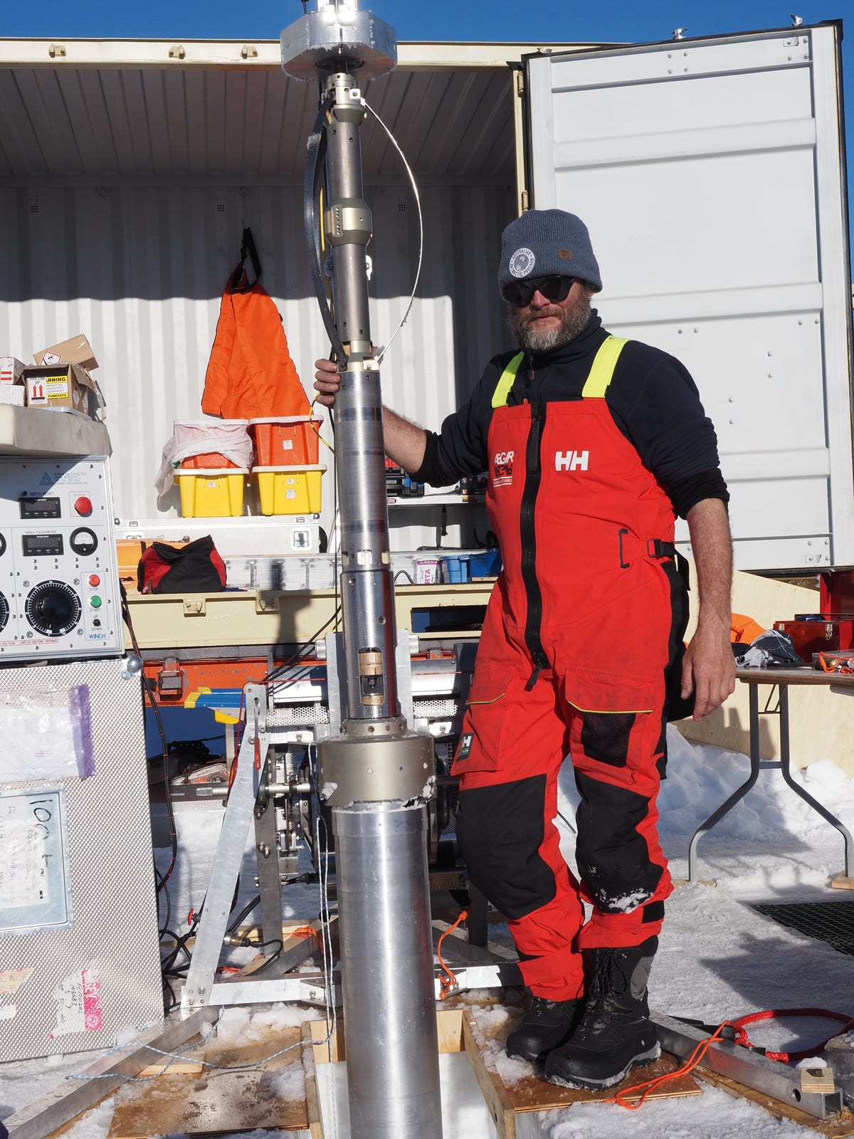 A man in red overalls and a black shirt, black sunglasses and a grey beanie stands with a large ice-coring drill.
