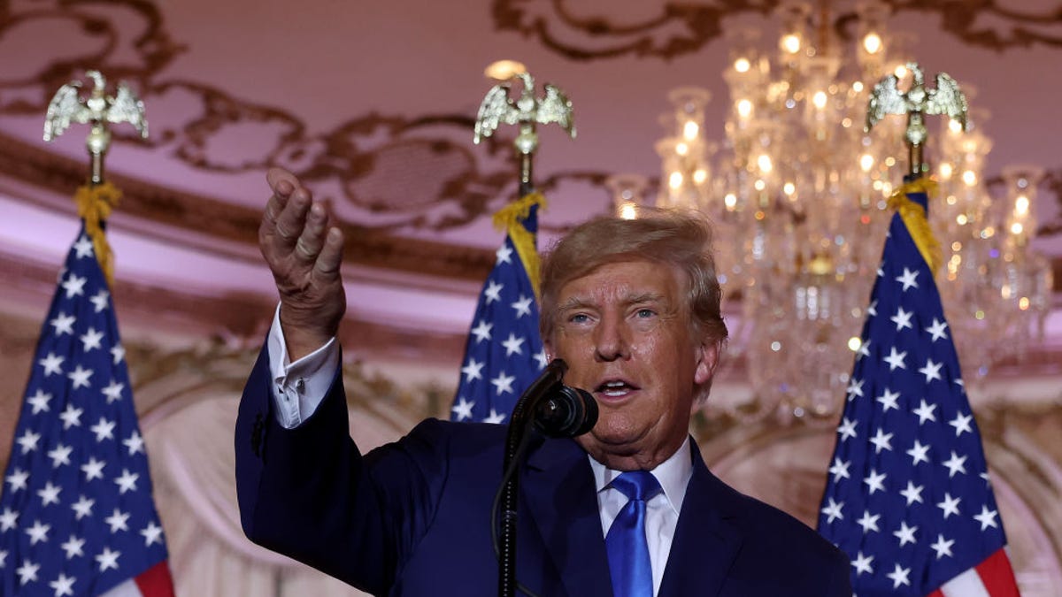 Former US President Donald Trump gestures with flags and a chandelier in the background