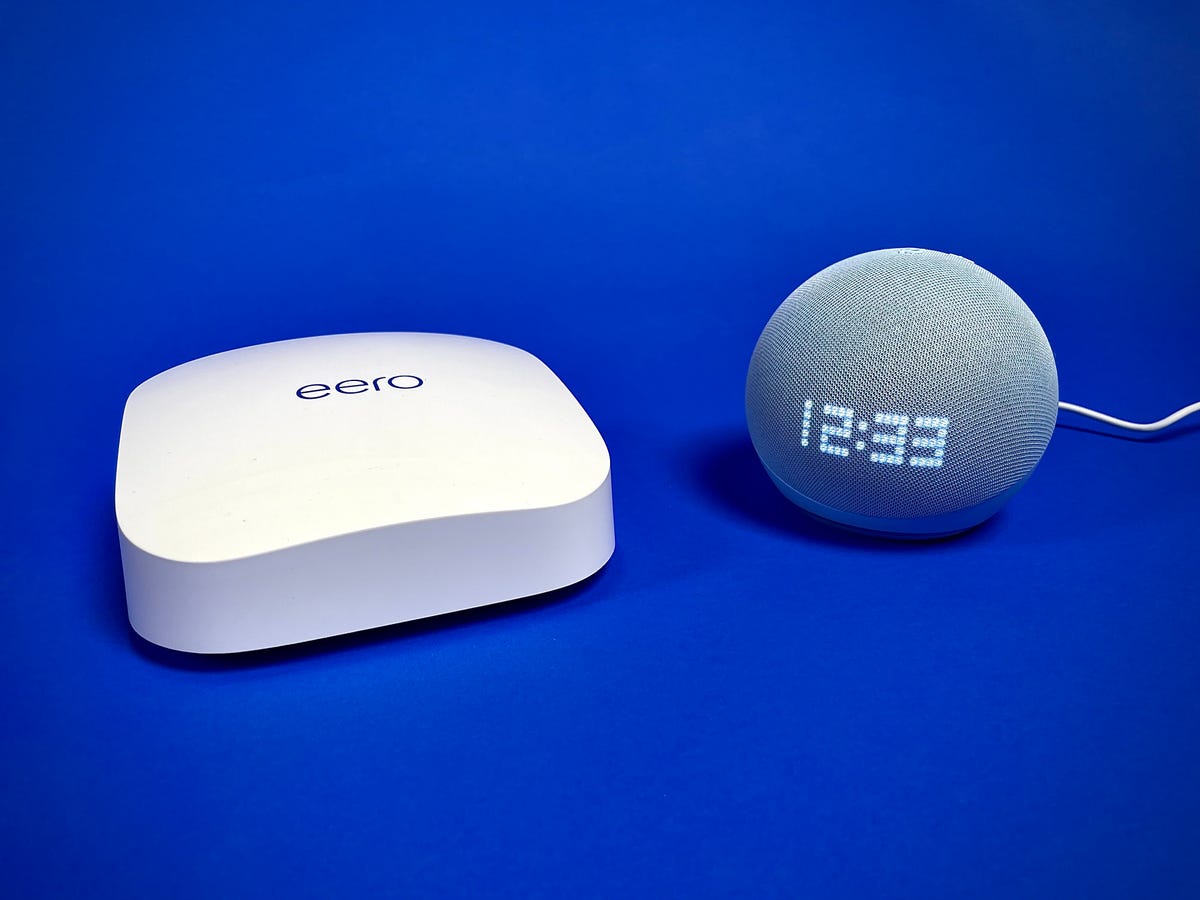 An Eero Pro 6E router sits beside the 5th-gen Amazon Echo Dot with Clock against a blue background. The new Echo Dots can double as Wi-Fi extenders for Eero's mesh networks.
