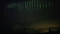 A still image captured by a camera on the Subaru Telescope in Hawaii shows the cloudy night sky with a curtain of individual downward streaks of green laser light.