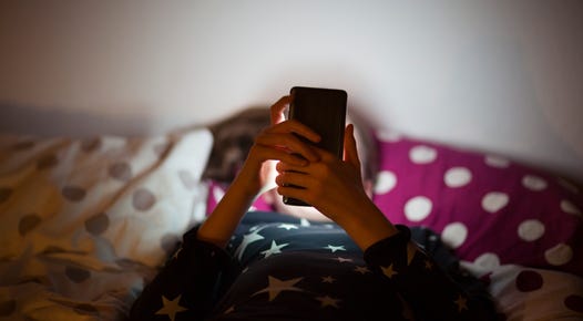A child lays in bed and stares up at her phone