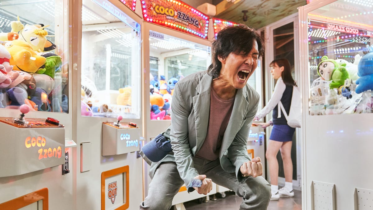 Lee Jung-jae, as Player 456 in Squid Game, screams in celebration in surrounded by brightly lit arcade games. 