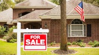 A brick house with an American flag flying and a for sale sign in the front yard.