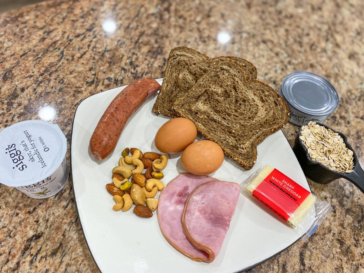 a spread of food containing yogurt, mixed nuts, sausage, ham, eggs, bread, cheese, oats, and tuna depicting 100 grams of protein