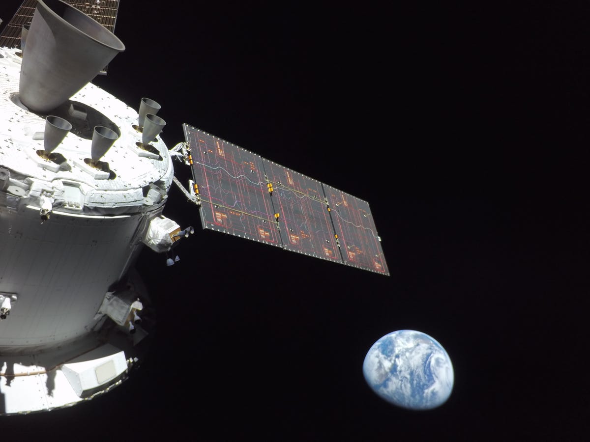 Часть космического корабля Орион в белом цвете с расширенной солнечной батареей и частичной Землей внизу, темное пространство вокруг.