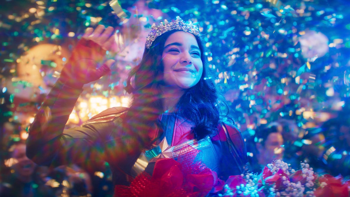 Trophy winner Kamala Khan smiles and waves in triumph as colorful confetti rains down on her.