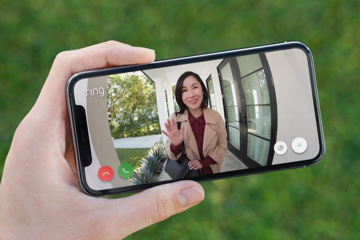 A smartphone in someone's hand showing a landscape view of the Ring Doorbell live view with a woman waving on the screen.