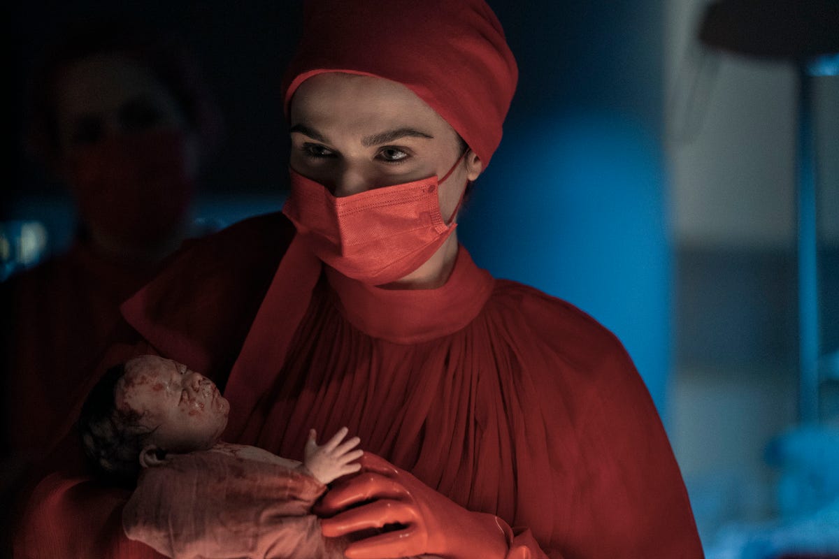 Rachel Weisz holding a newborn baby, wearing red surgical garments