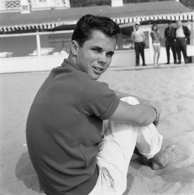 Actor Tony Dow sitting on a beach, looking over his shoulder