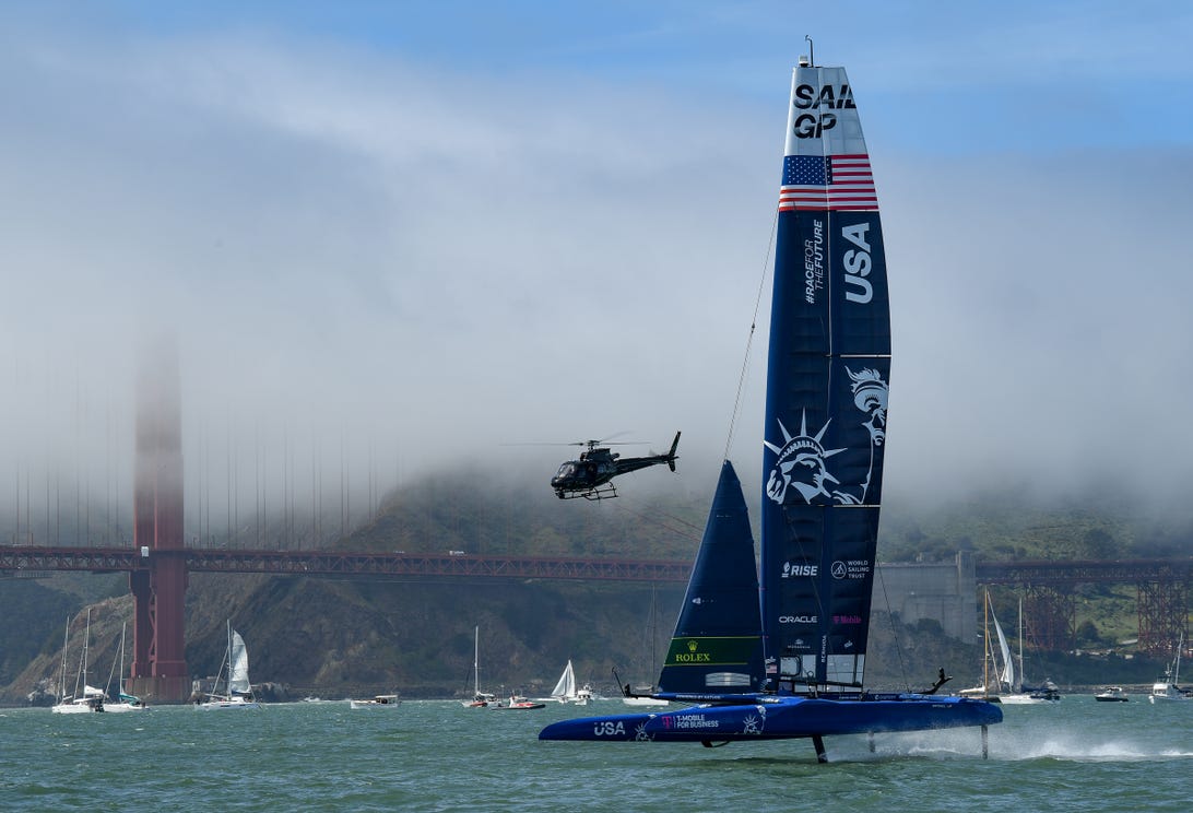 The Team USA F50 sailboat sails in front of the Golden Gate Bridge as a helicopter hovers nearby.