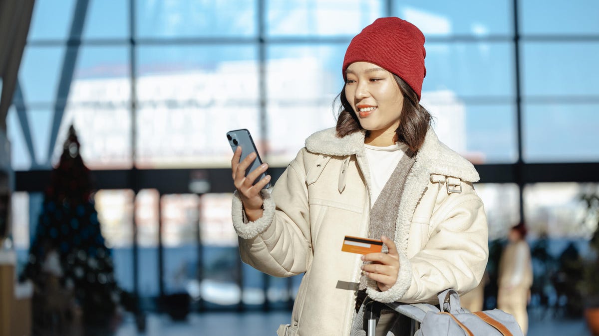 A woman holds her phone and a bank card while standing in an airport.