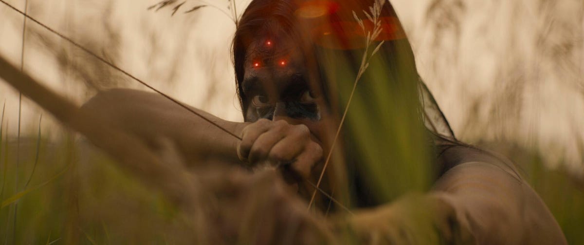 Red dots glare on woman's forehead as she holds a bow and arrow while hiding in reeds