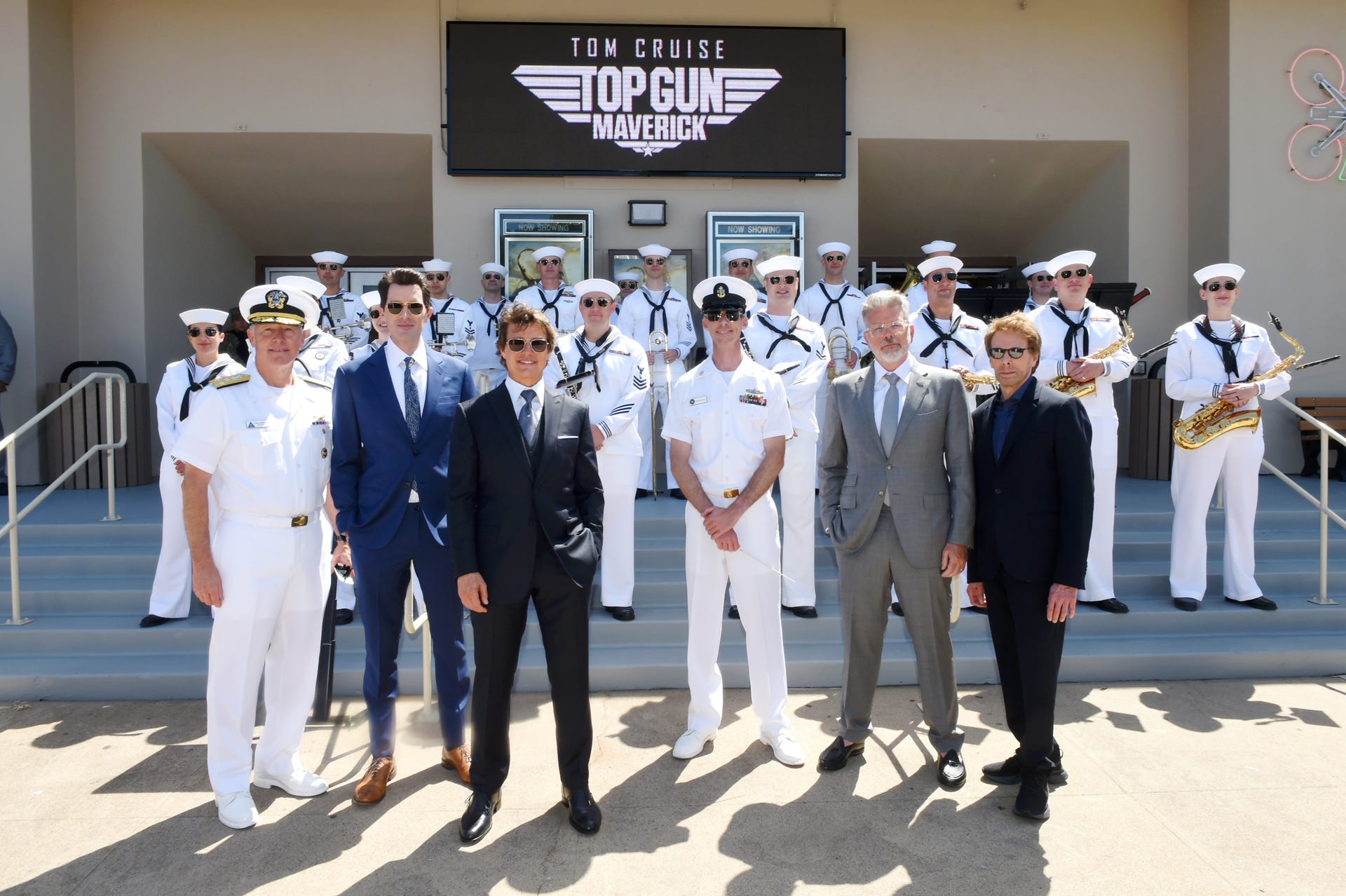 Tom Cruise, Joseph Kosinski, Christopher McQuarrie, Jerry Bruckheimer wear suits and line up with US Navy band members and top brass in gleaming white uniforms under a Top Gun banner.