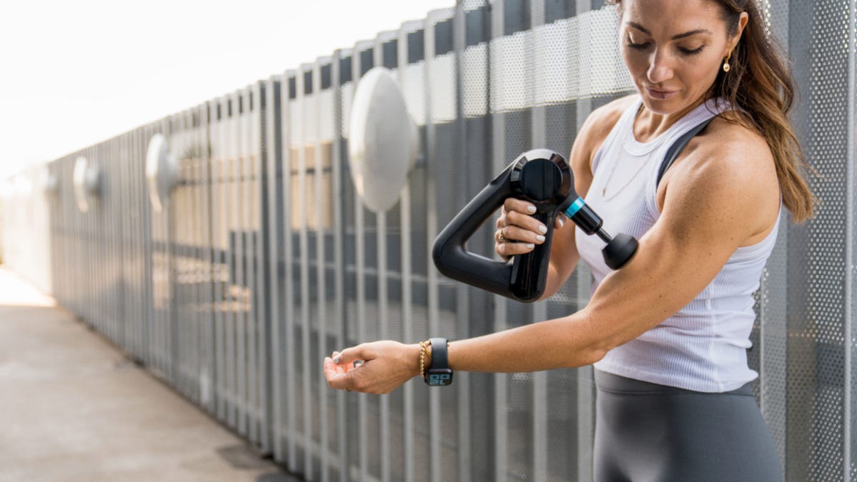 A woman in workout clothes using a Theragun on her arm