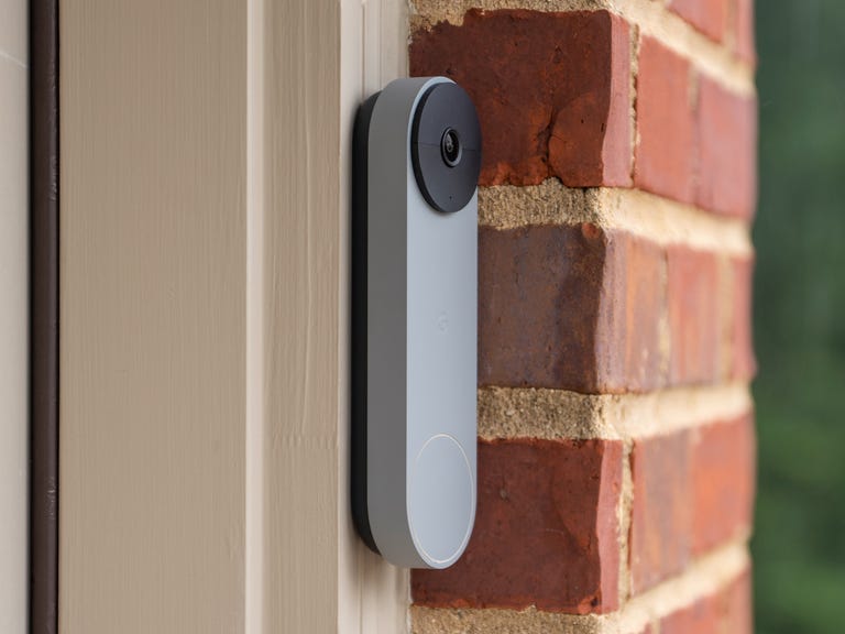 The Nest Doorbell (battery) mounted on an off-white door frame, next to a brick wall and greenery.