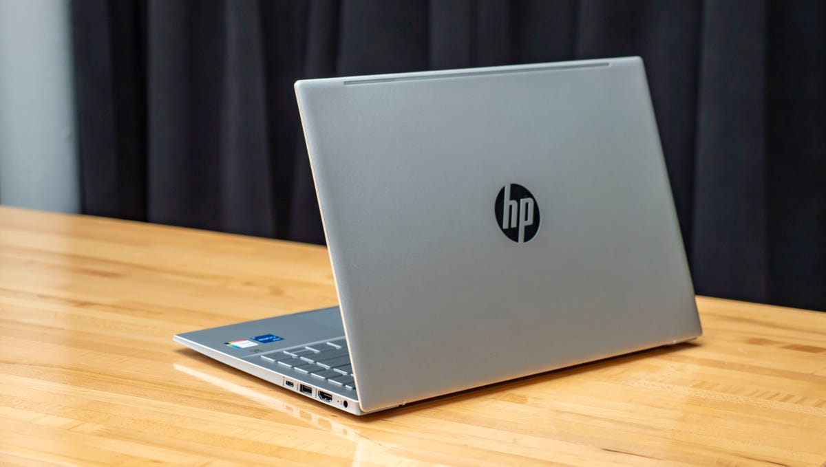 HP Pavilion 14 laptop open and showing the lid with an HP logo and facing to the left on a wood table with a black curtain behind it.