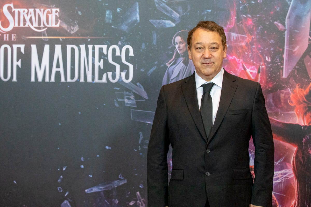 Director Sam Raimi stands in front of a large Doctor Strange in the Multiverse of Madness poster