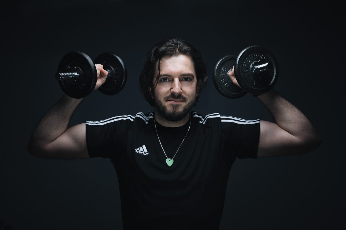 A man in a black Adidas T-shirt holds dumbbells by his head