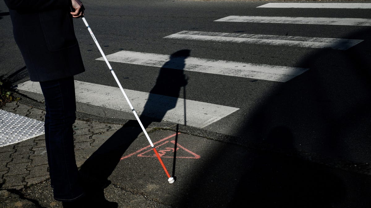 Photo of a visually impaired person with a cane, standing next to a crosswalk.