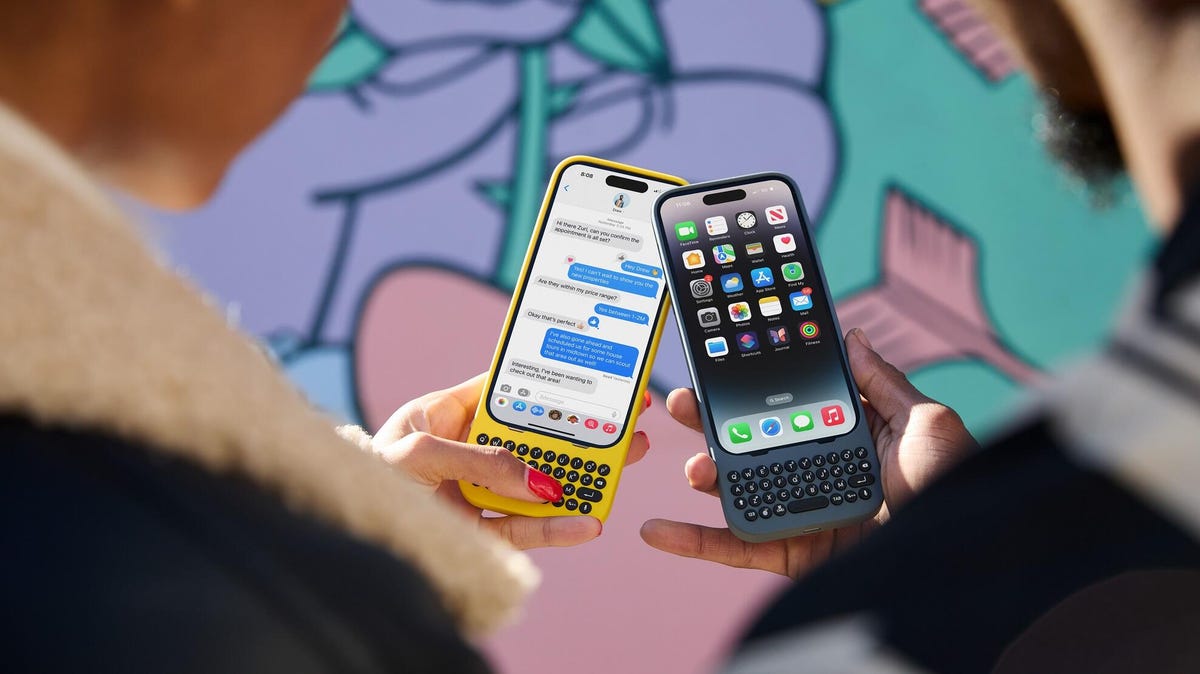 Two iPhones are held next to each other, each with a Clicks keyboard case around it -- one bright yellow, one a darker smoke gray.