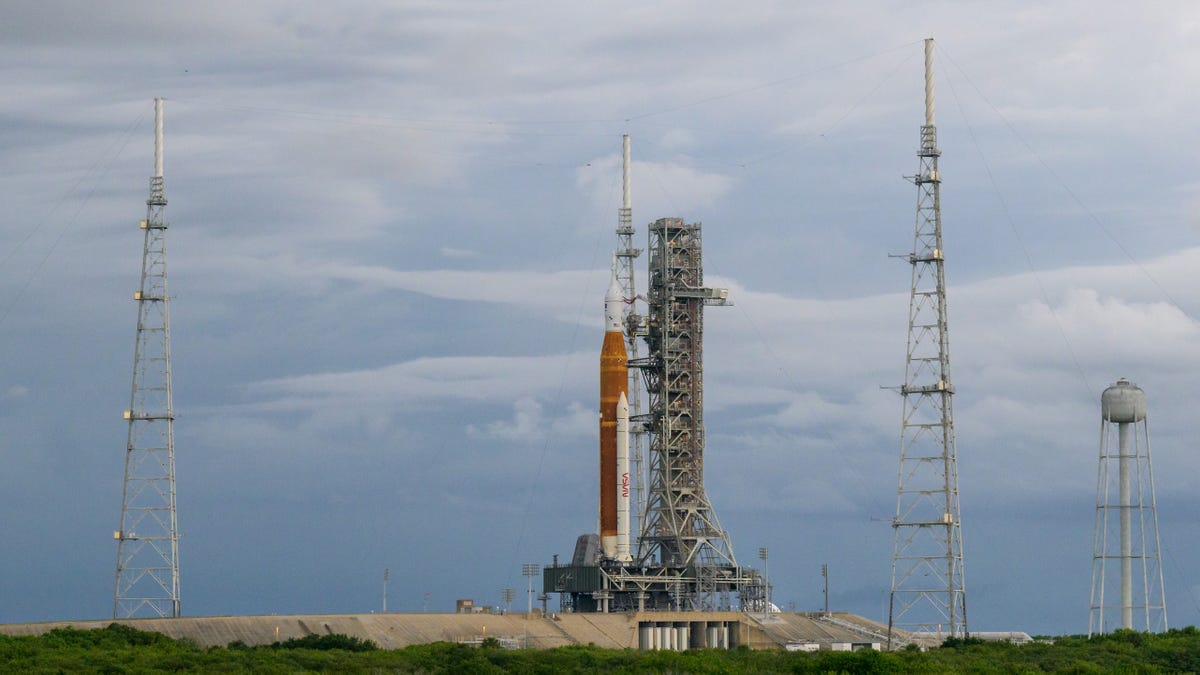 The orange and white Space Launch System stands on the launchpad next to a green tower. Lightning towers are visible to the left and right of the launchpad