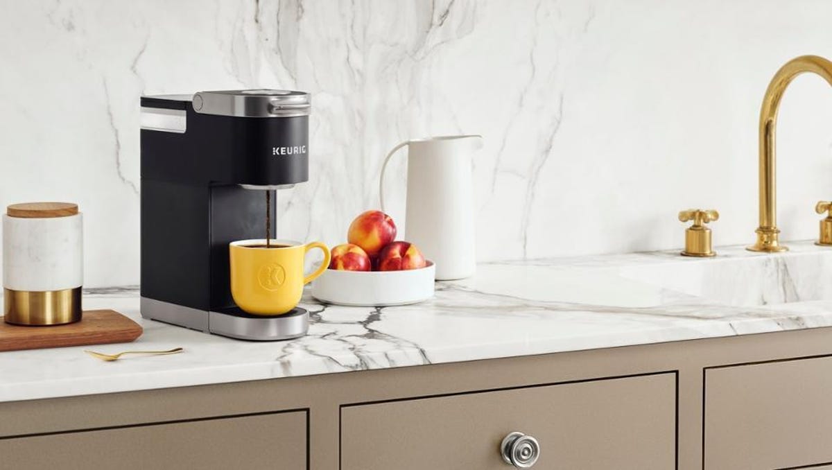A black Keurig K-Slim on a white marble countertop with a bowl of fruit and a cup of coffee.