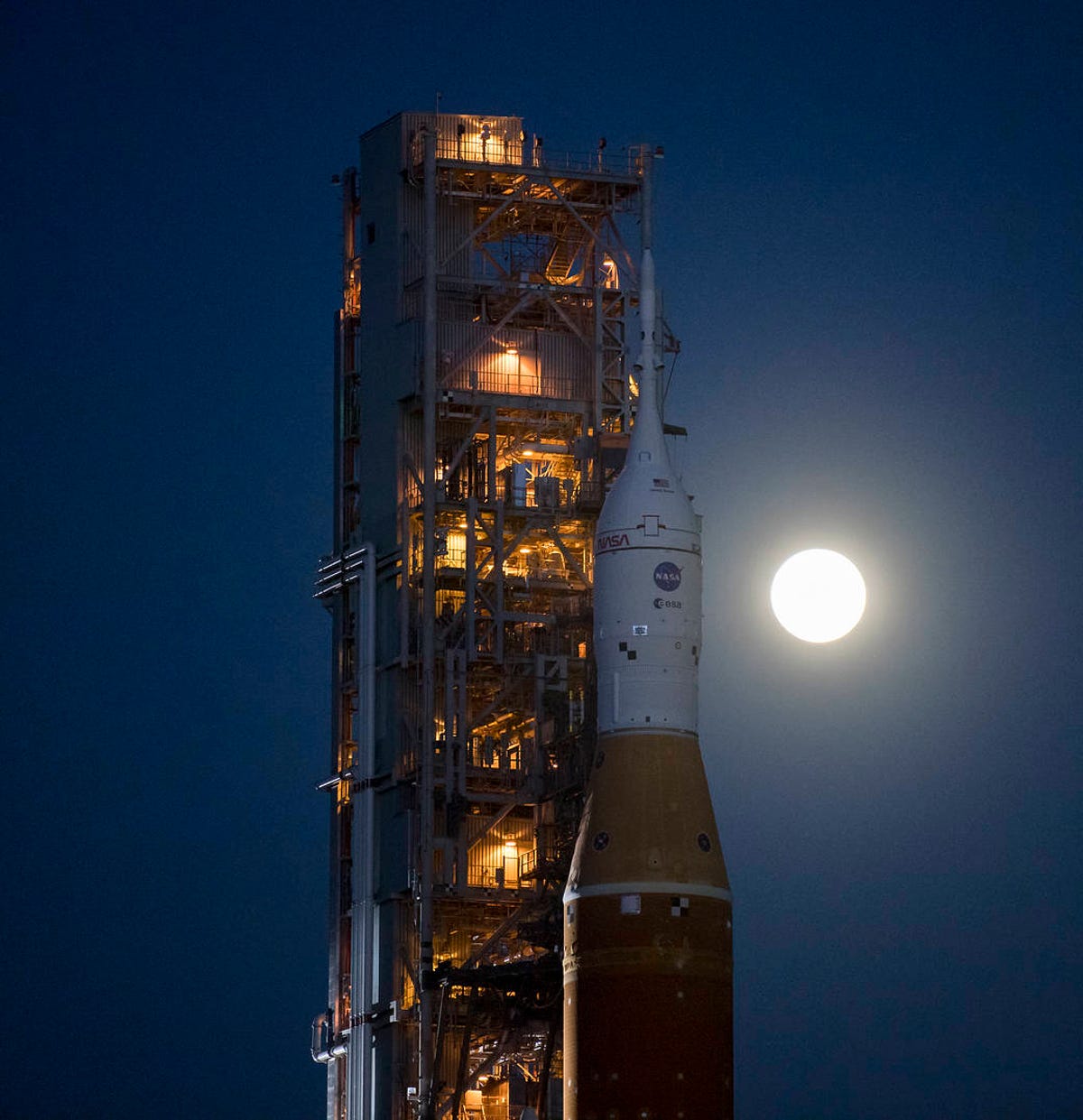 It's dusk, and the top of NASA's orange SLS rocket attached to the white Orion spacecraft is seen with a brilliant, glowing moon in the background.