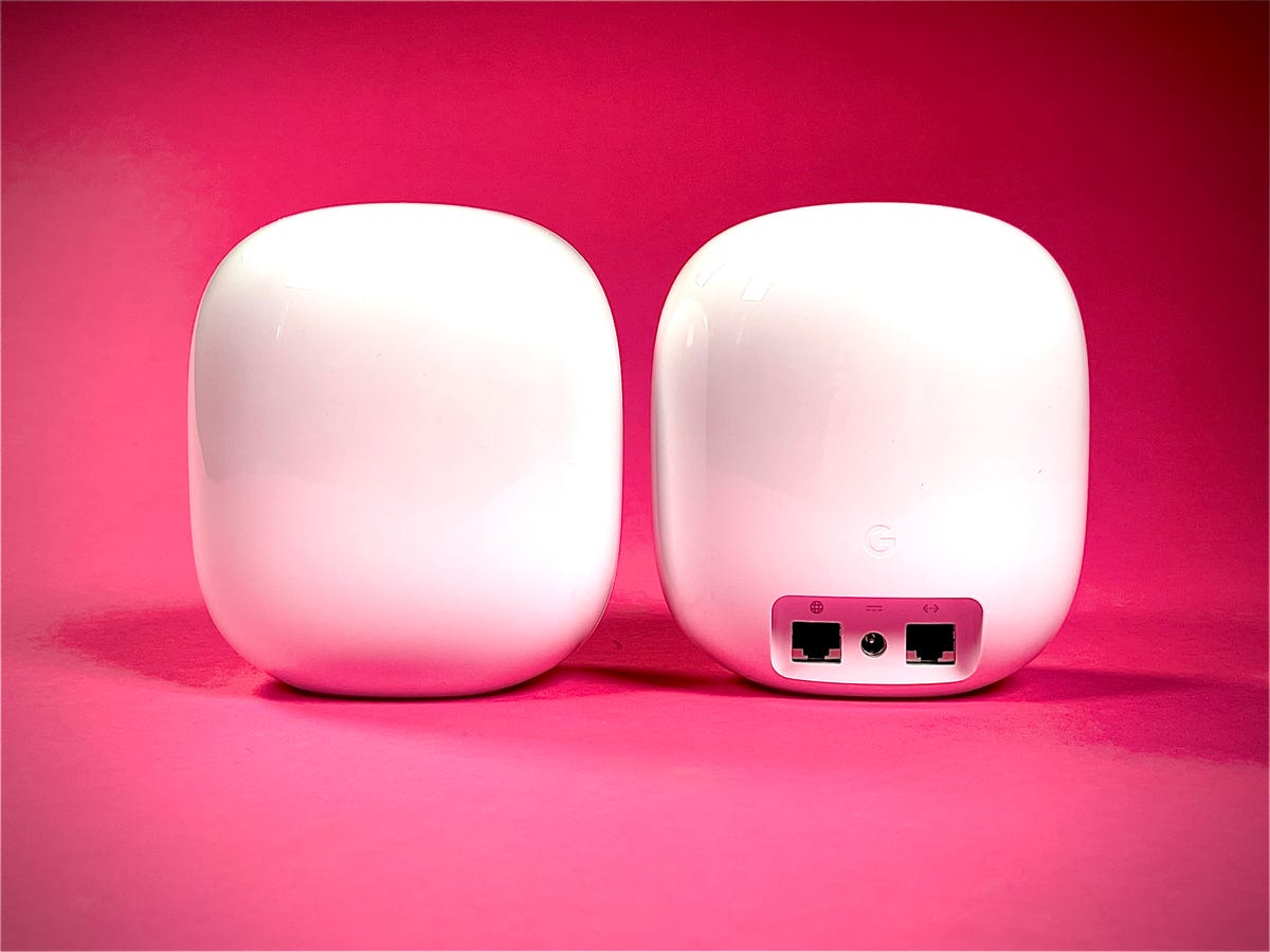 Two Nest Wifi Pro mesh routers sit side by side against a pink background, one facing us the other turned to show the ports in the back. Each device has an A/C power jack and two gigabit Ethernet ports.