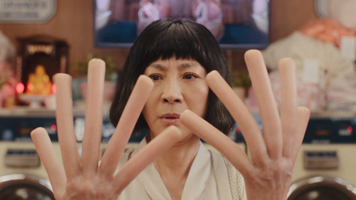 Michelle Yeoh stars as Evelyn in Everything Everywhere All at Once. This still shows her with a shortish black bob haircut and a white shirt holding up her hands with fingers shaped like hot dogs.