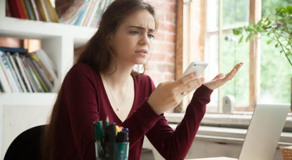 Frustrated young woman looking at her phone while sitting at her home desk as her laptop is open.