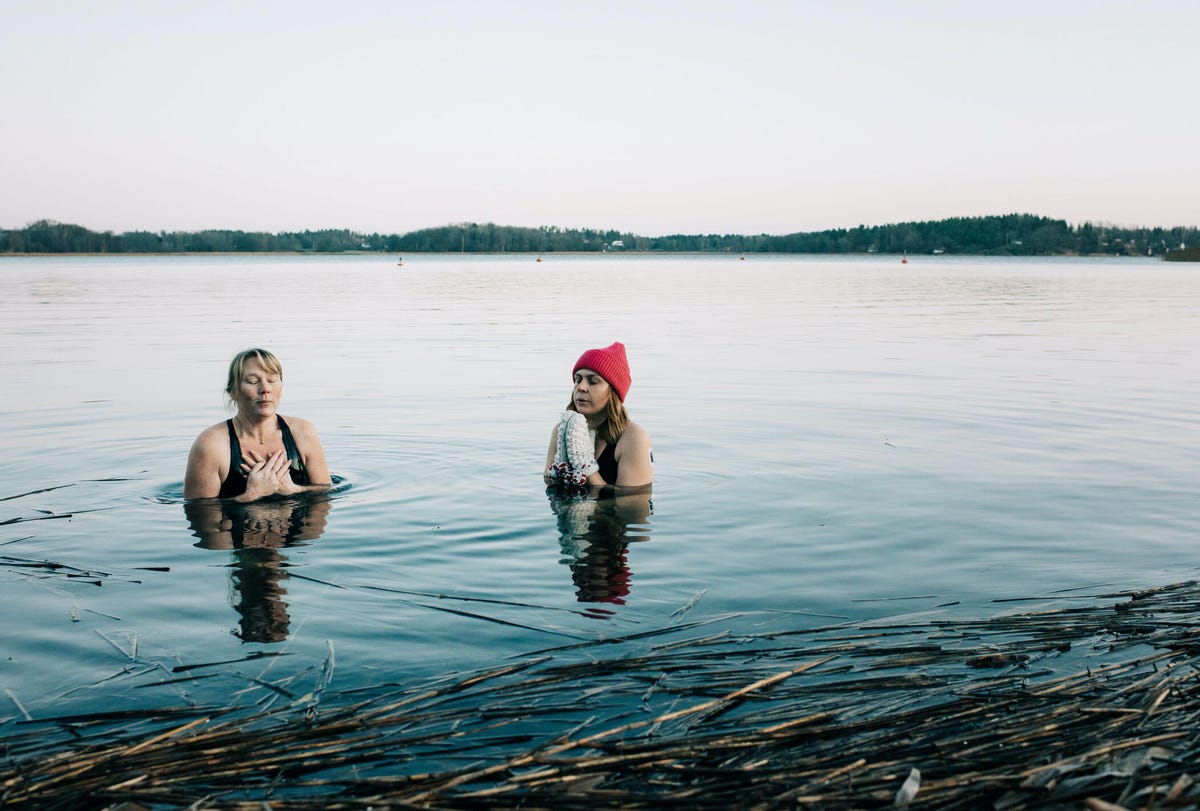 Two woman in bathing suits in a lake. One wears a red knit cap and white mittens.