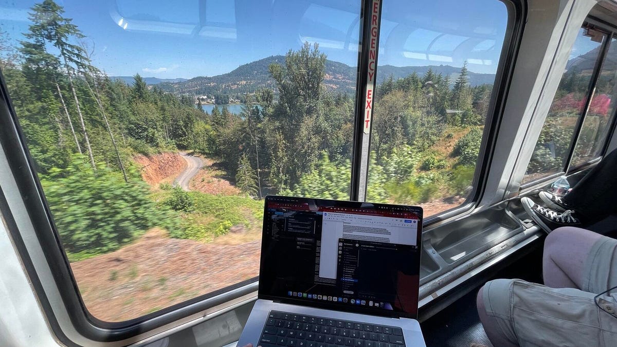 The reporter's laptop in front of the lounge car's ample windows, behind which a gorgeous spread of tall, verdant trees atop brown underbrush and a clear blue lake behind it visible through the treeline.