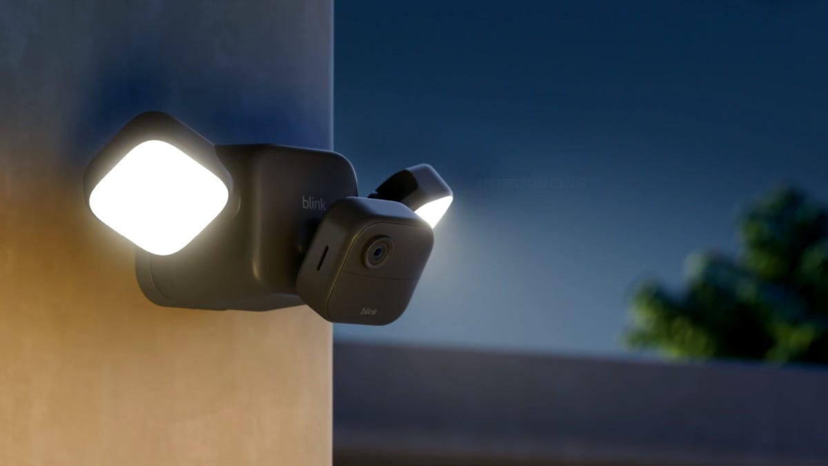 A Blink Outdoor 4 Floodlight Cam attached to the side of a home at dusk casts its 700 lumens of light into the yard.