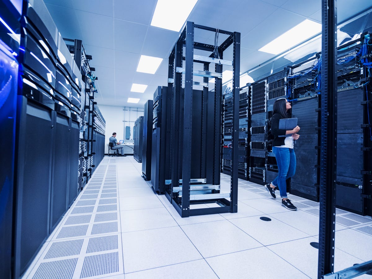 Web server room. Someone in the background is working at a desk while a woman walks among the servers