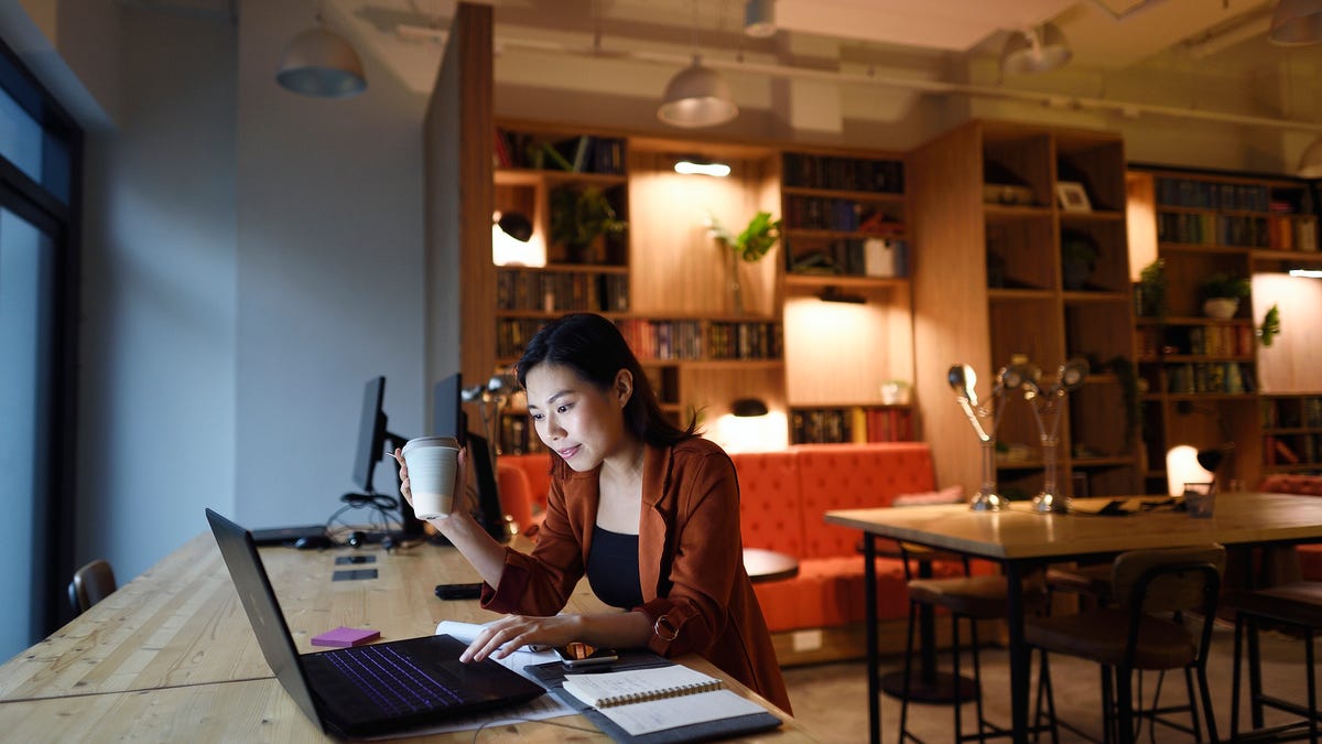 Unidentified woman is holding a coffee cup in her right hand as she glances down at her computer screen in her office.