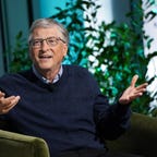 Bill Gates wearing a blue sweater and seated in a chair, gesturing with his hands