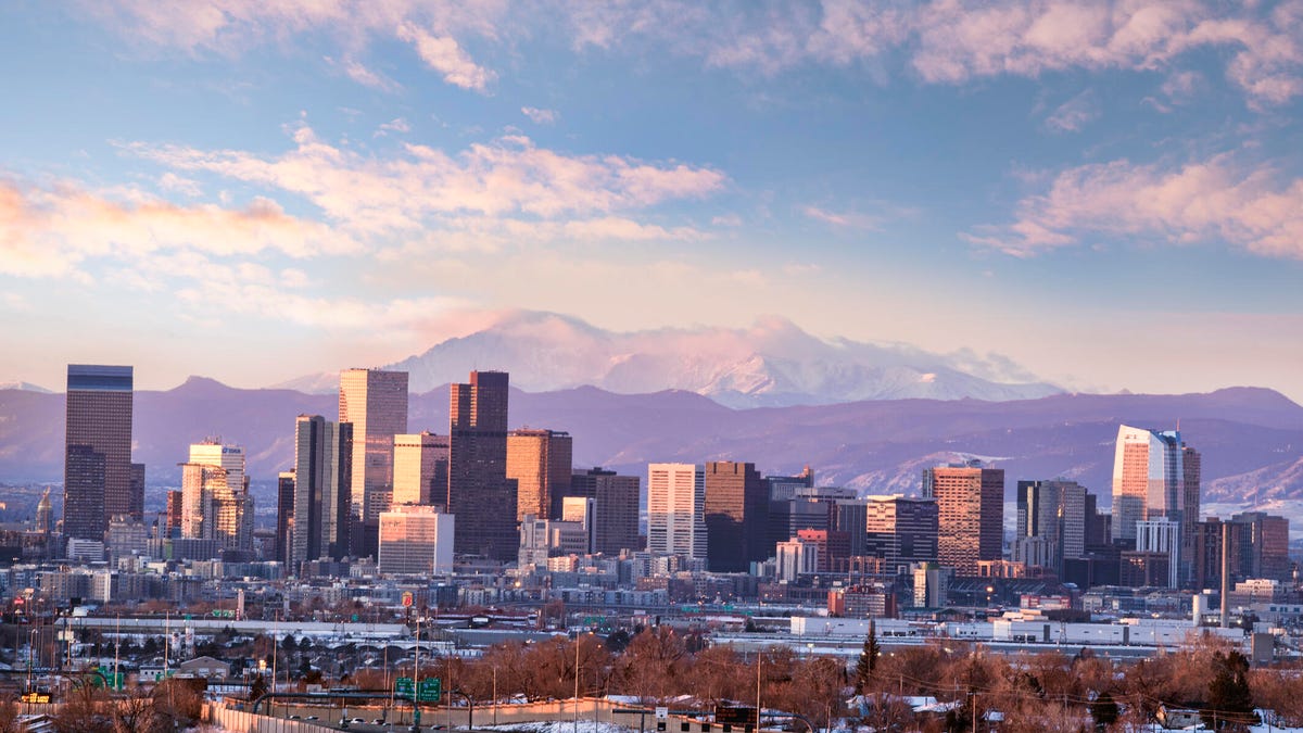 Denver skyline in front of a snowy mountain under a mostly sunny sky.