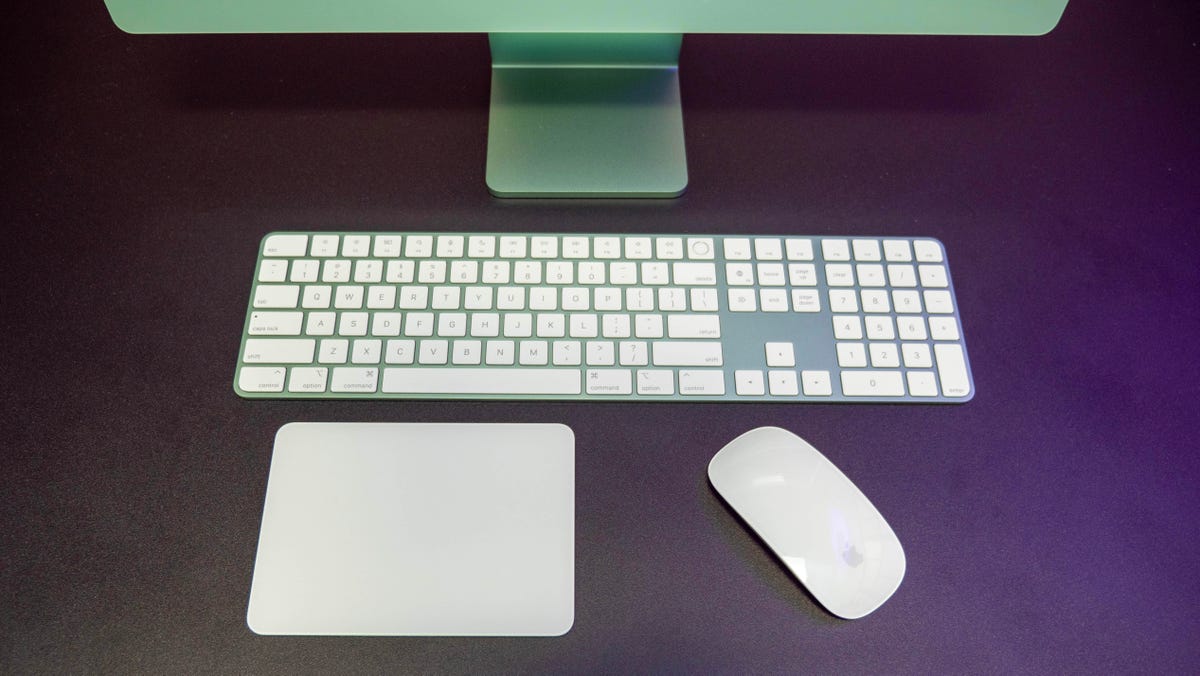 Apple iMac M3 Magic Keyboard, Magic Trackpad and Magic Mouse in green on a black desk in front of the base of the iMac.