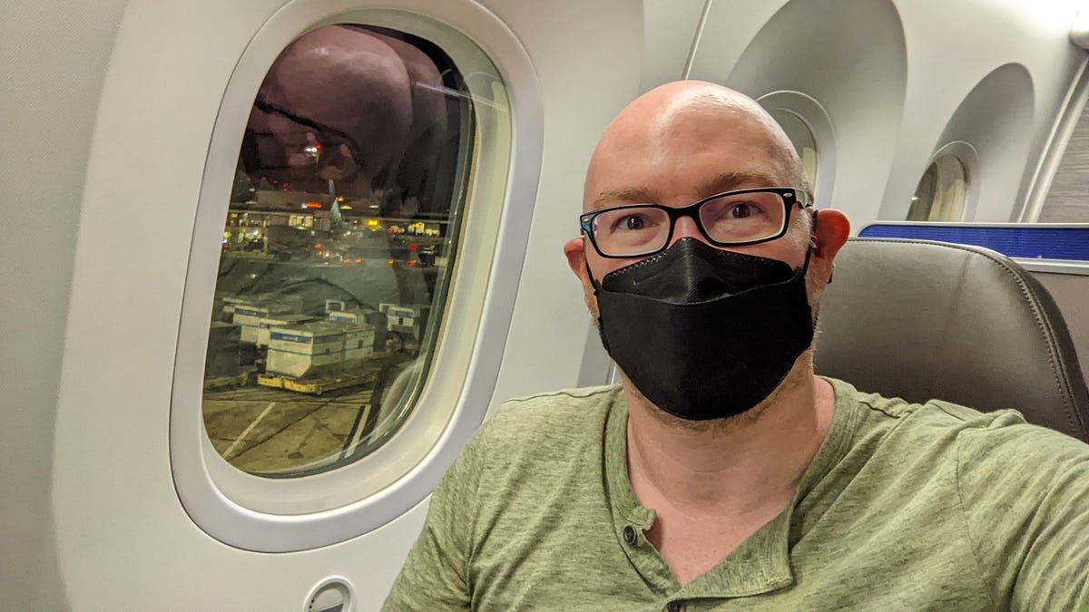 A bald man sits in front of a large airliner window. He wears a black mask.