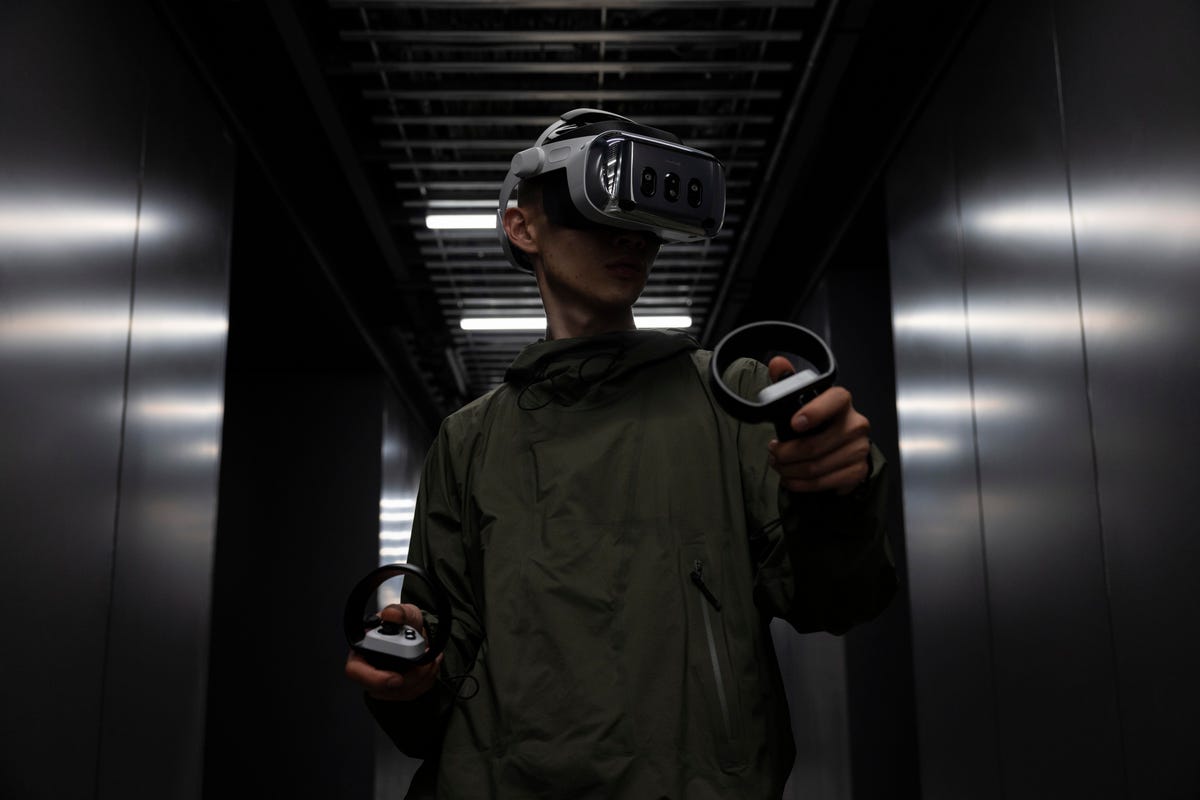 A person in a dark room wearing a Varjo XR-4 VR headset, and holding controllers|1200x800.390625
