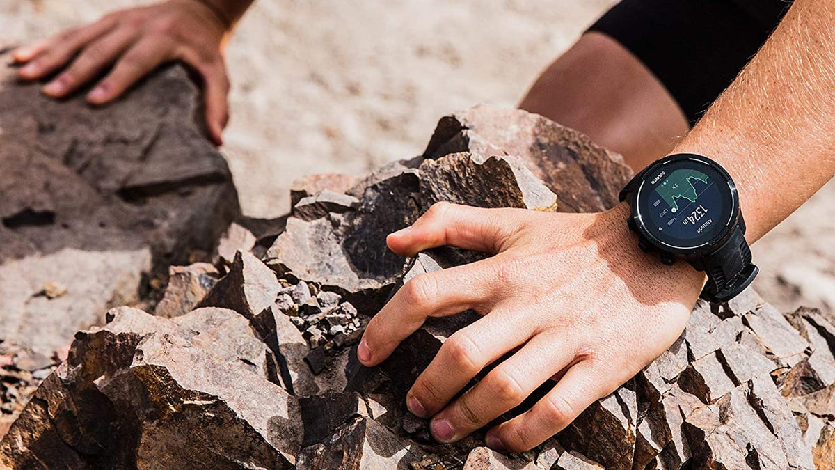 The hands of a person are placed on rugged, rocky terrain and the Suunto 9 sports watch rests on the left wrist.