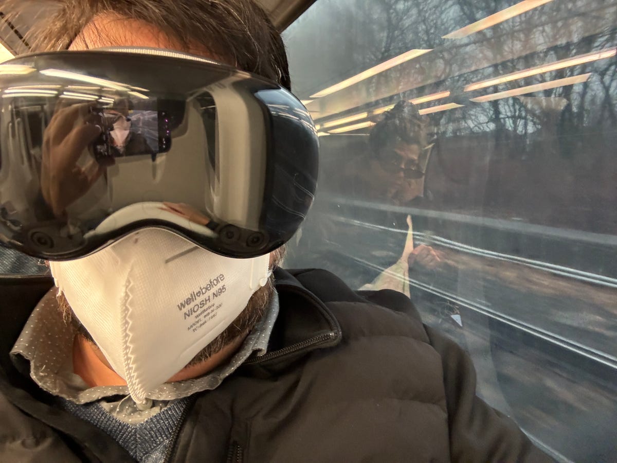 Someone wearing an Apple Vision Pro on a train with a mask on.