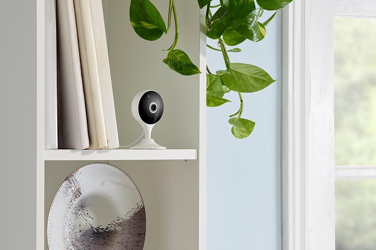 The Lorex security camera sitting on a white shelf in a white room beside a plant and various minimalistic decor.
